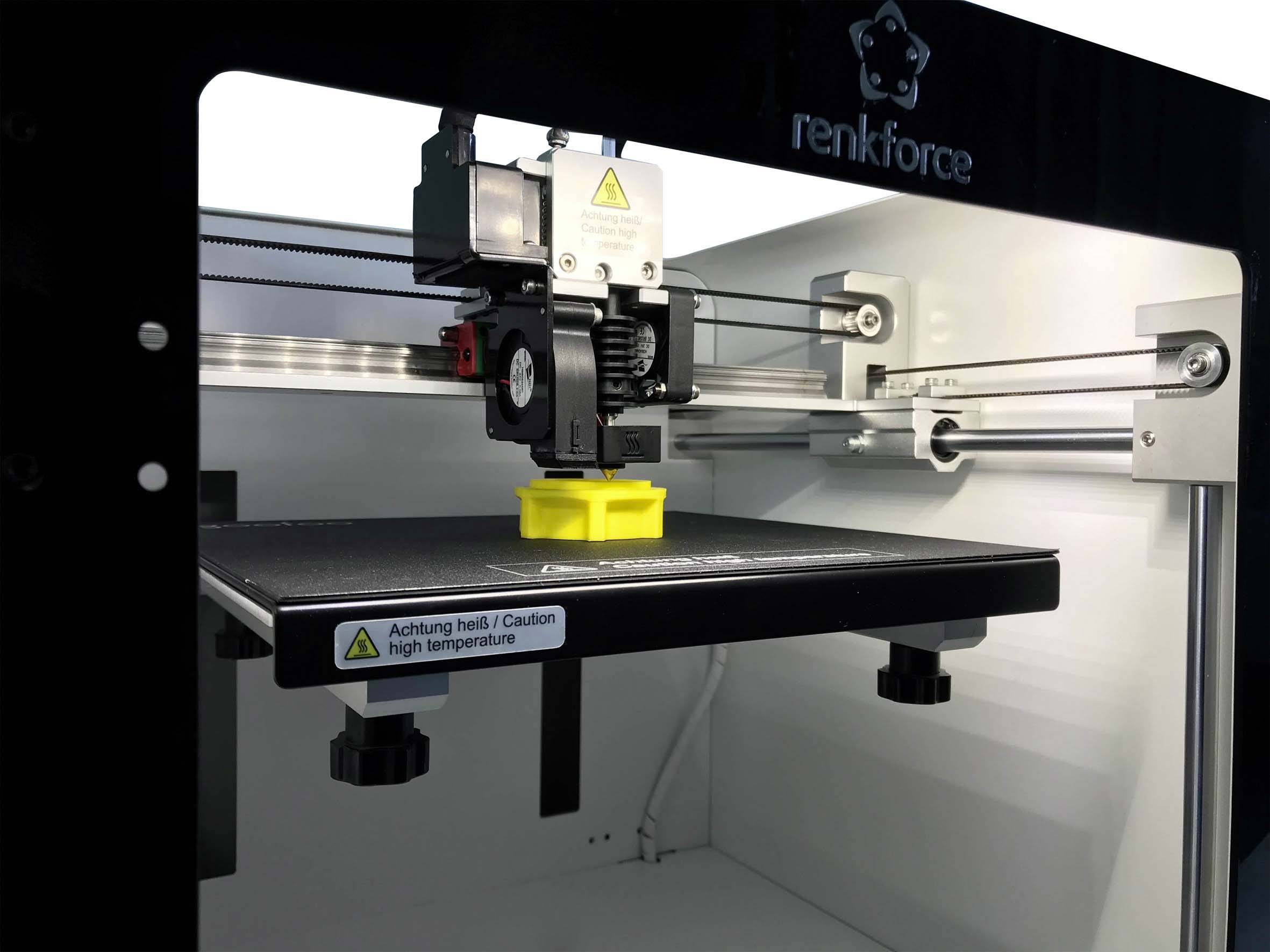 Een 3D-printer drukt een geel object. Waarschuwingsbord op het platform: 'Pas op, heet / Let op hoge temperatuur'.