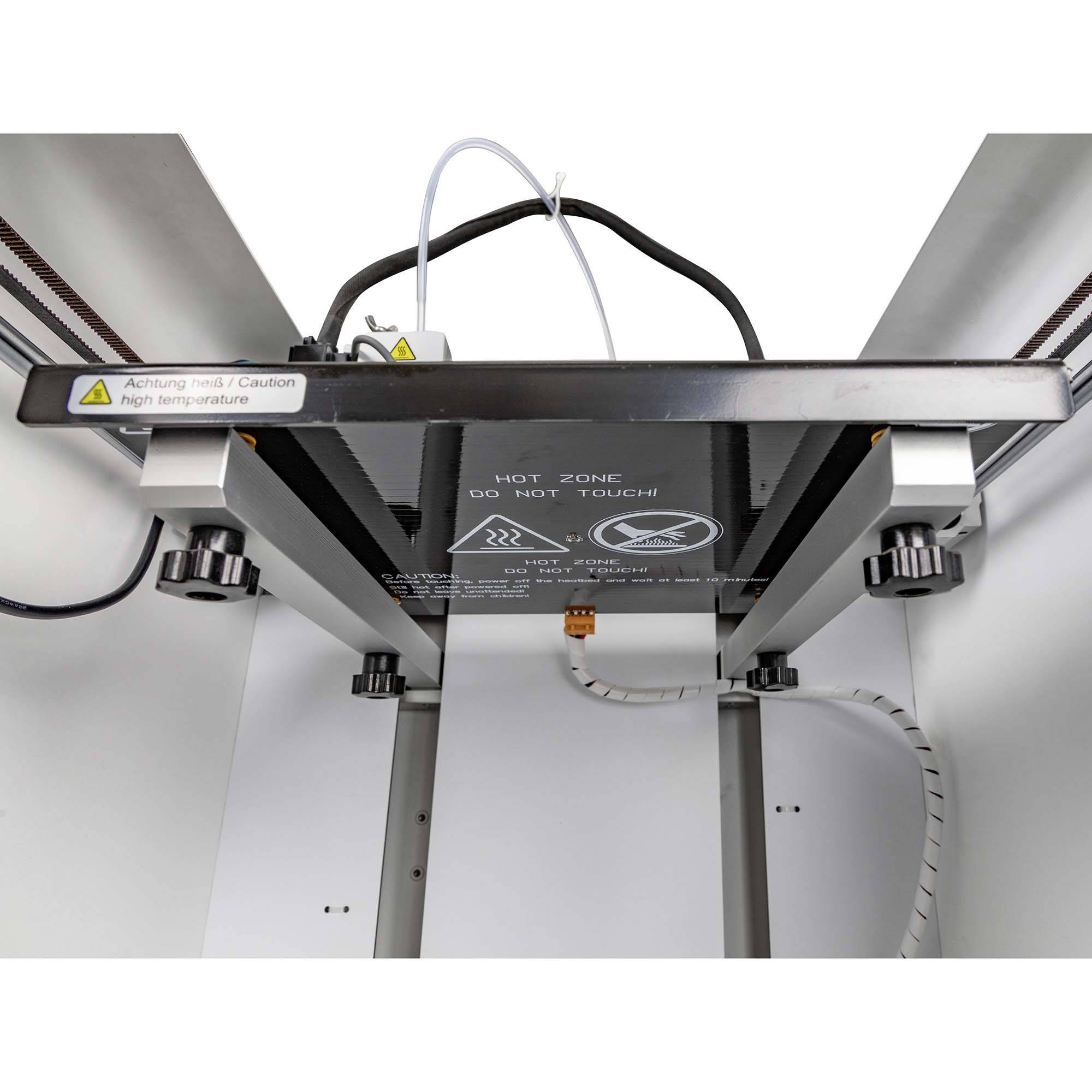 3D-printer gezien van onderen met een waarschuwingsbord: 'Hete Zone - Niet aanraken!'. Kabels en mechanische onderdelen zichtbaar.