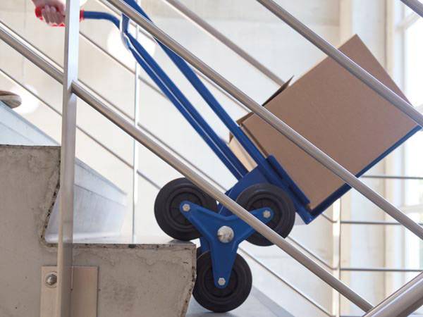 Een hand trekt een blauwe trapklimmer die een kartonnen doos de trap op transporteert.