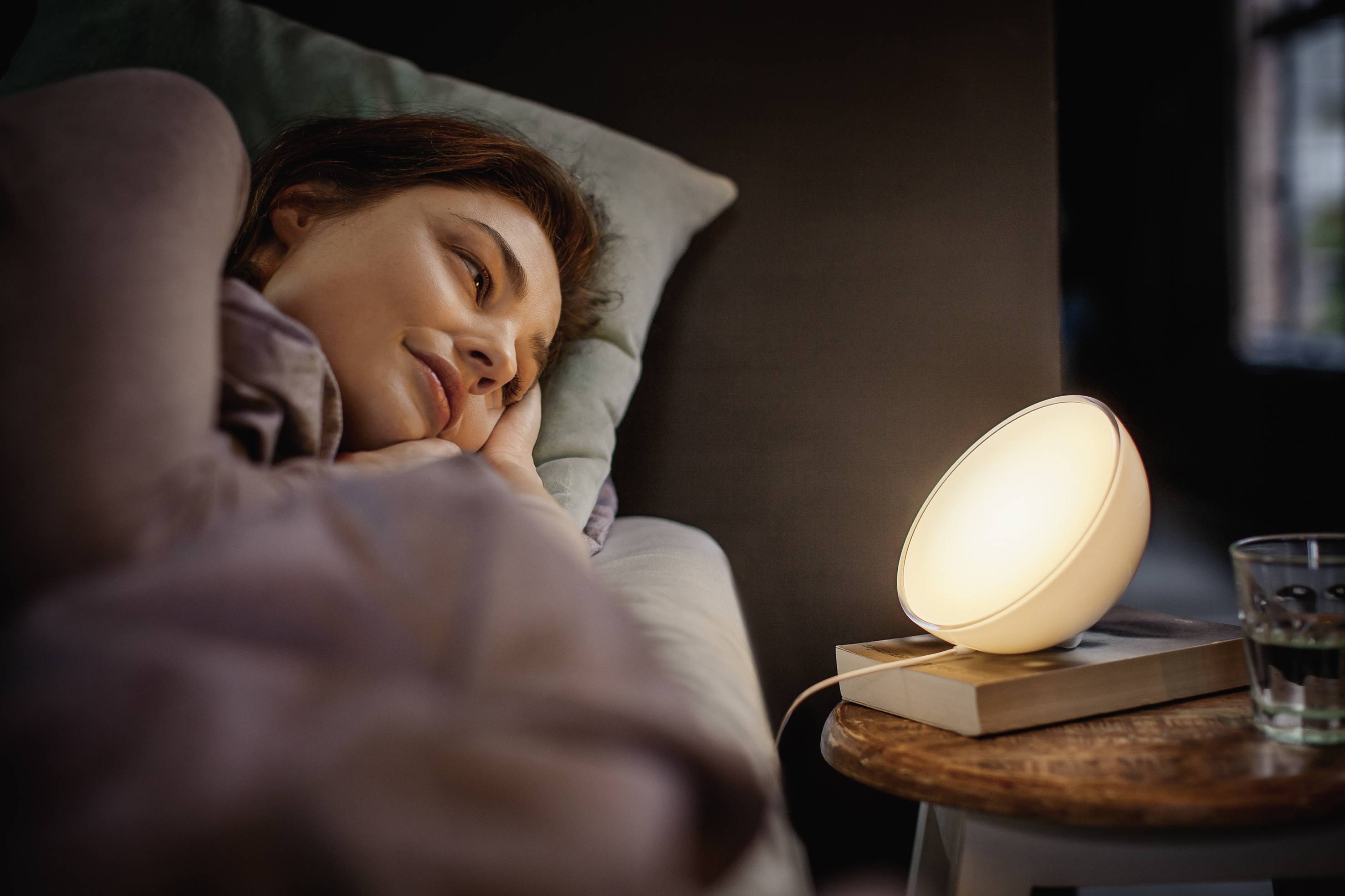 Vrouw ligt in bed, kijkt naar een verlichte lamp op het nachtkastje. Sfeervolle verlichting creëert een ontspannen sfeer.