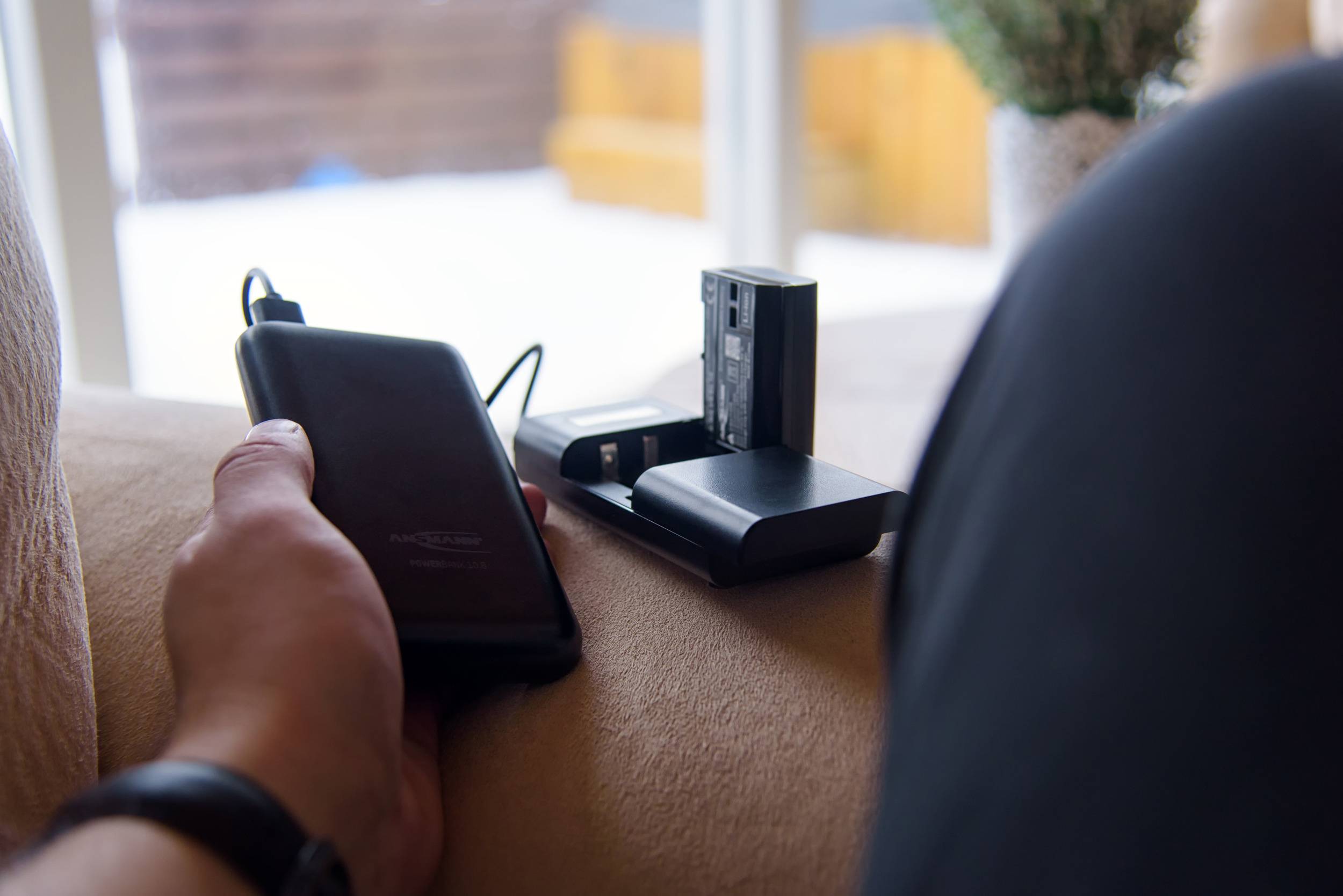Een persoon houdt een powerbank en twee oplaadbare batterijen op een oplader, met op de achtergrond wazig een raam.