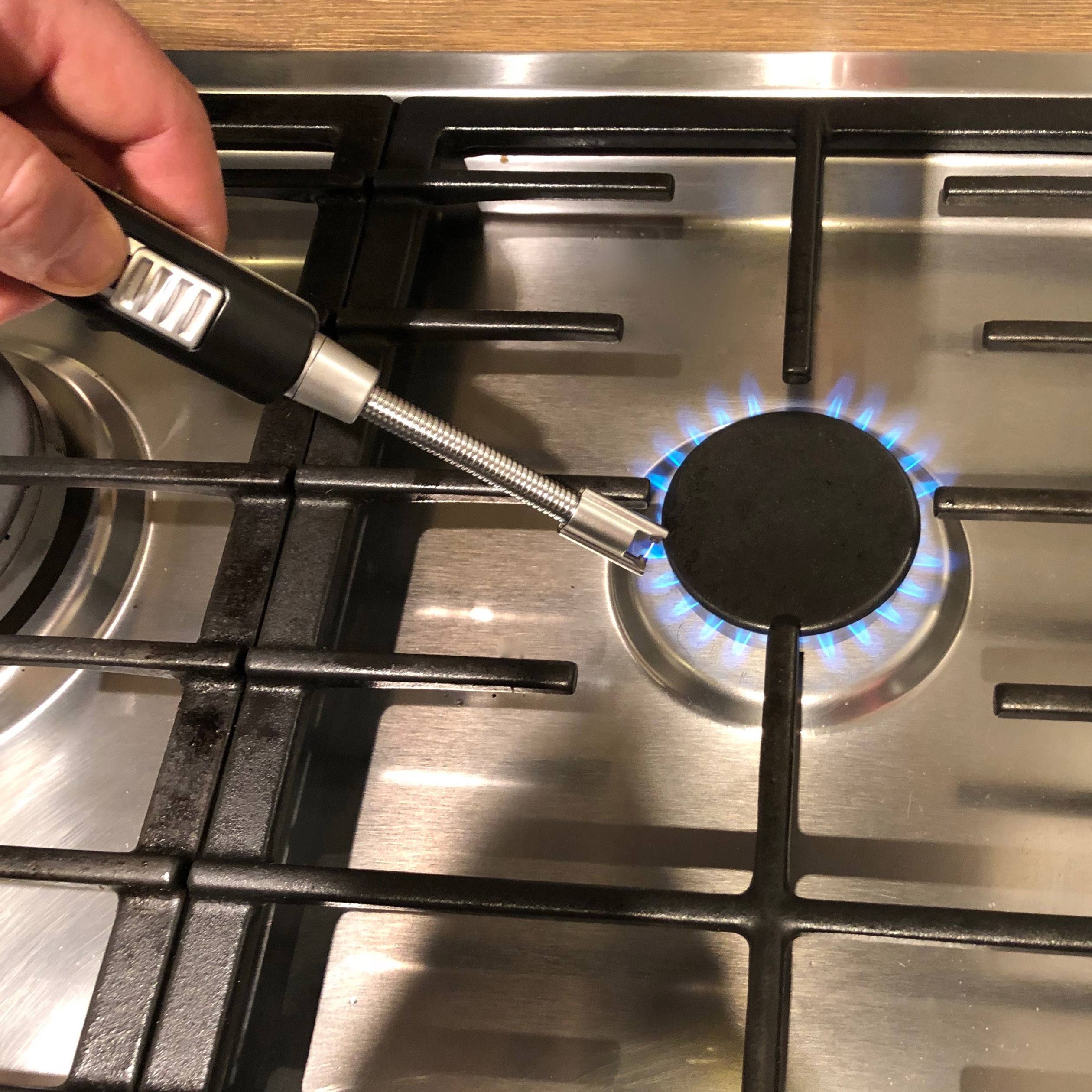 Een hand houdt een vuurpistool met lange steel, gericht op een ingeschakeld gasfornuis; de vlam brandt blauw.