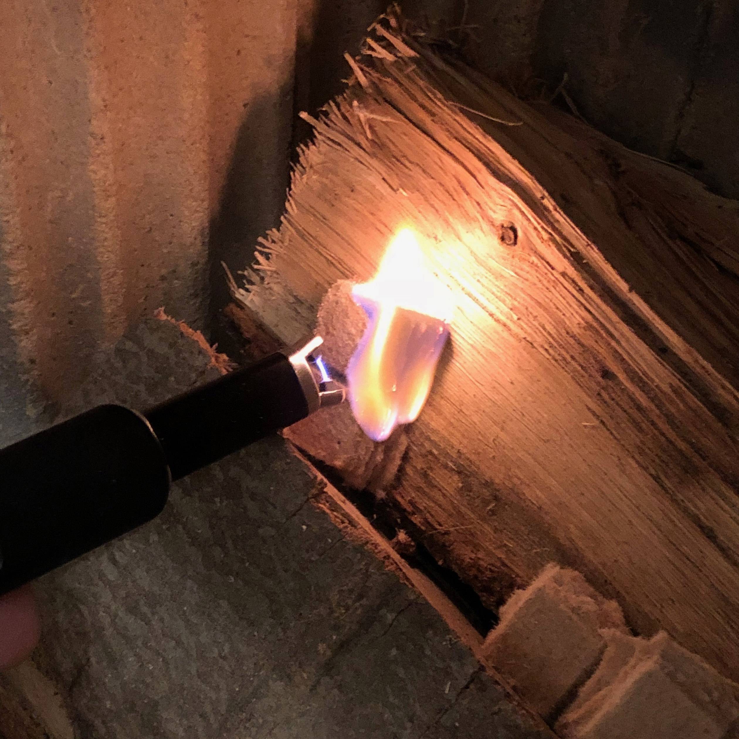 Een elektrische aansteker steekt een stuk hout aan. De vlam is helder en het hout begint te branden.