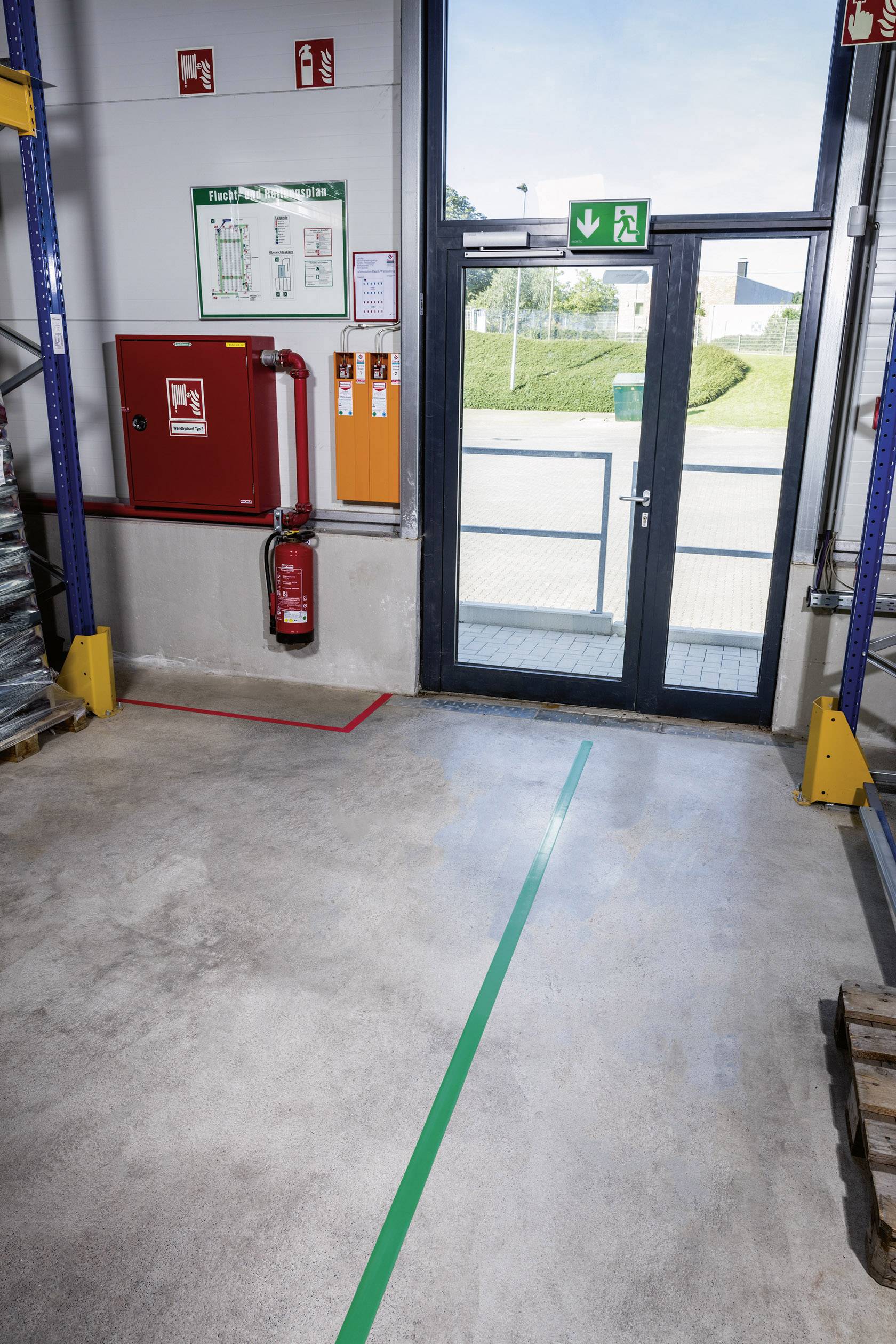 Een uitgang in een magazijn met glazen deuren, daarboven een groen nooduitgangsbord. Brandblusser en vluchtroute-instructies zijn aan de muur bevestigd.