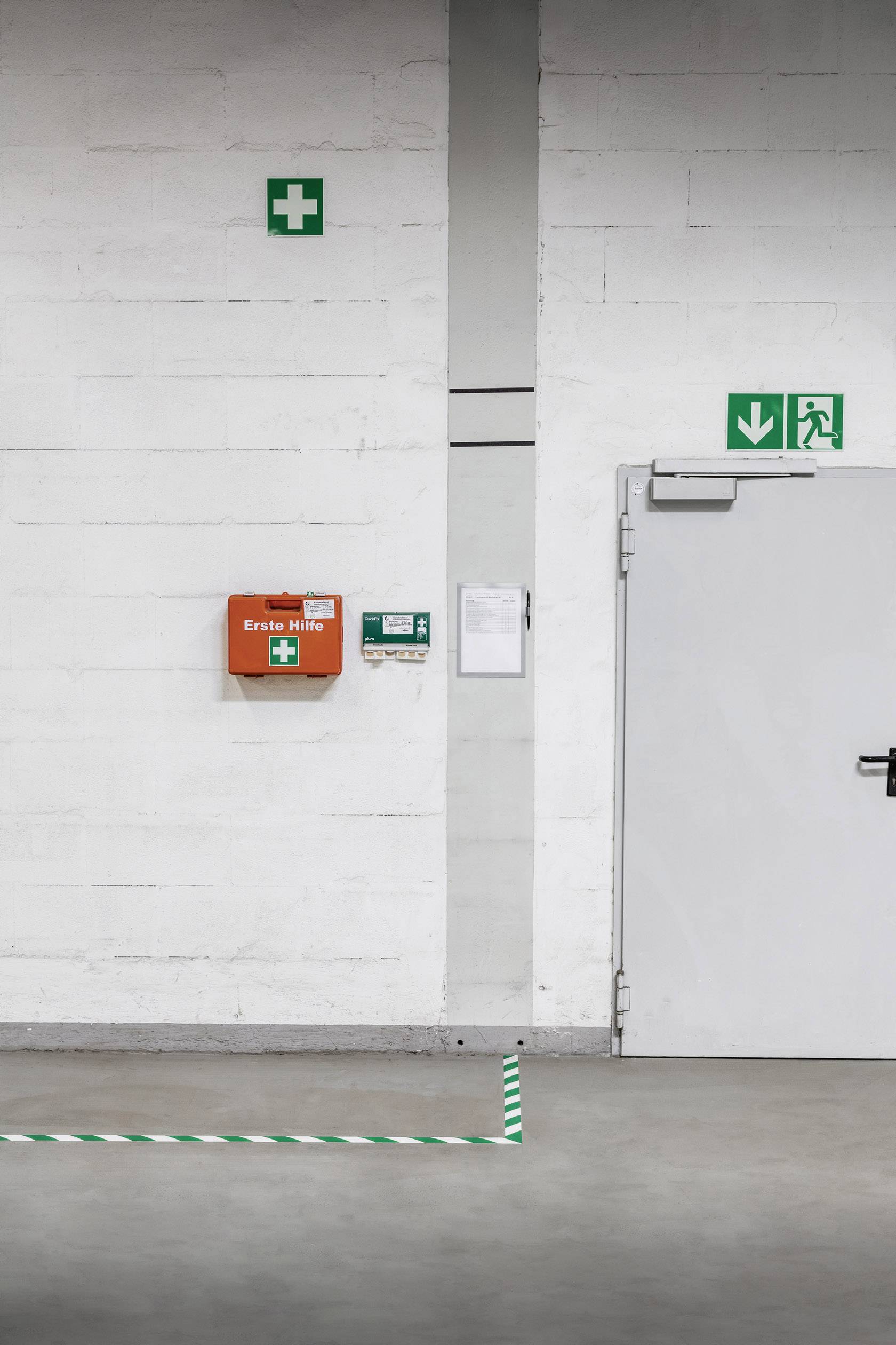 Een muur met een EHBO-doos, een pijlteken voor nooduitgang en een deur in een industriële ruimte.
