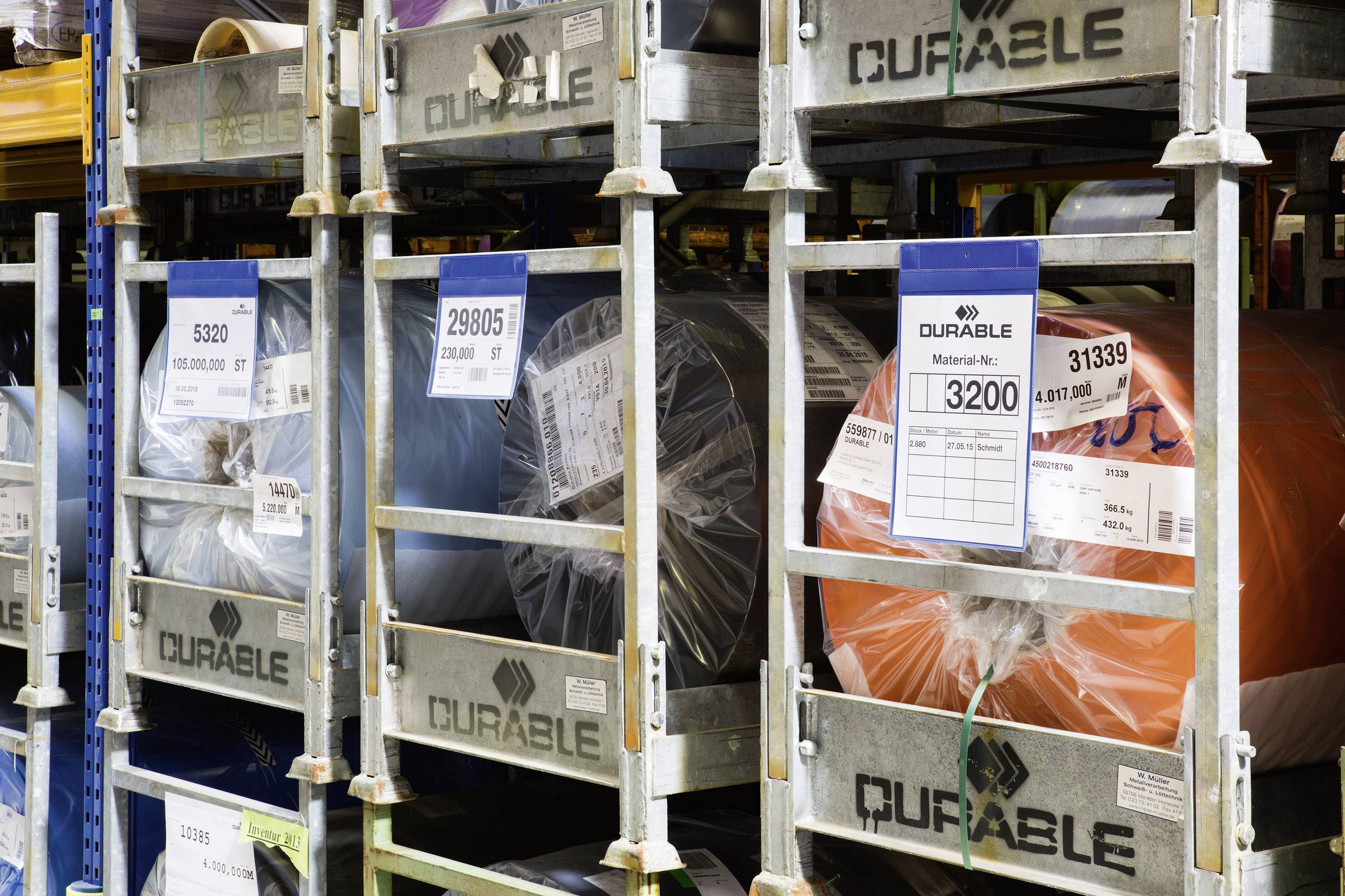 Opslagrekken met grote kunststof rollen in verschillende kleuren. Etiketten geven materiaalnummers en gewichten aan.