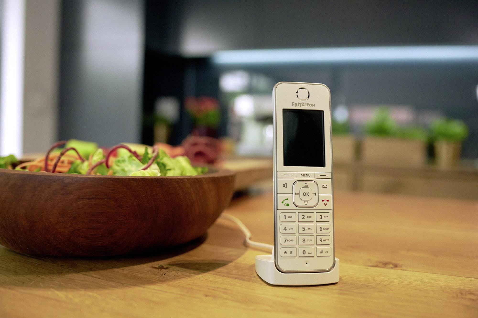 Een draadloze telefoon staat op een tafel naast een kom met salade. Op de achtergrond zijn wazig keukenobjecten zichtbaar.