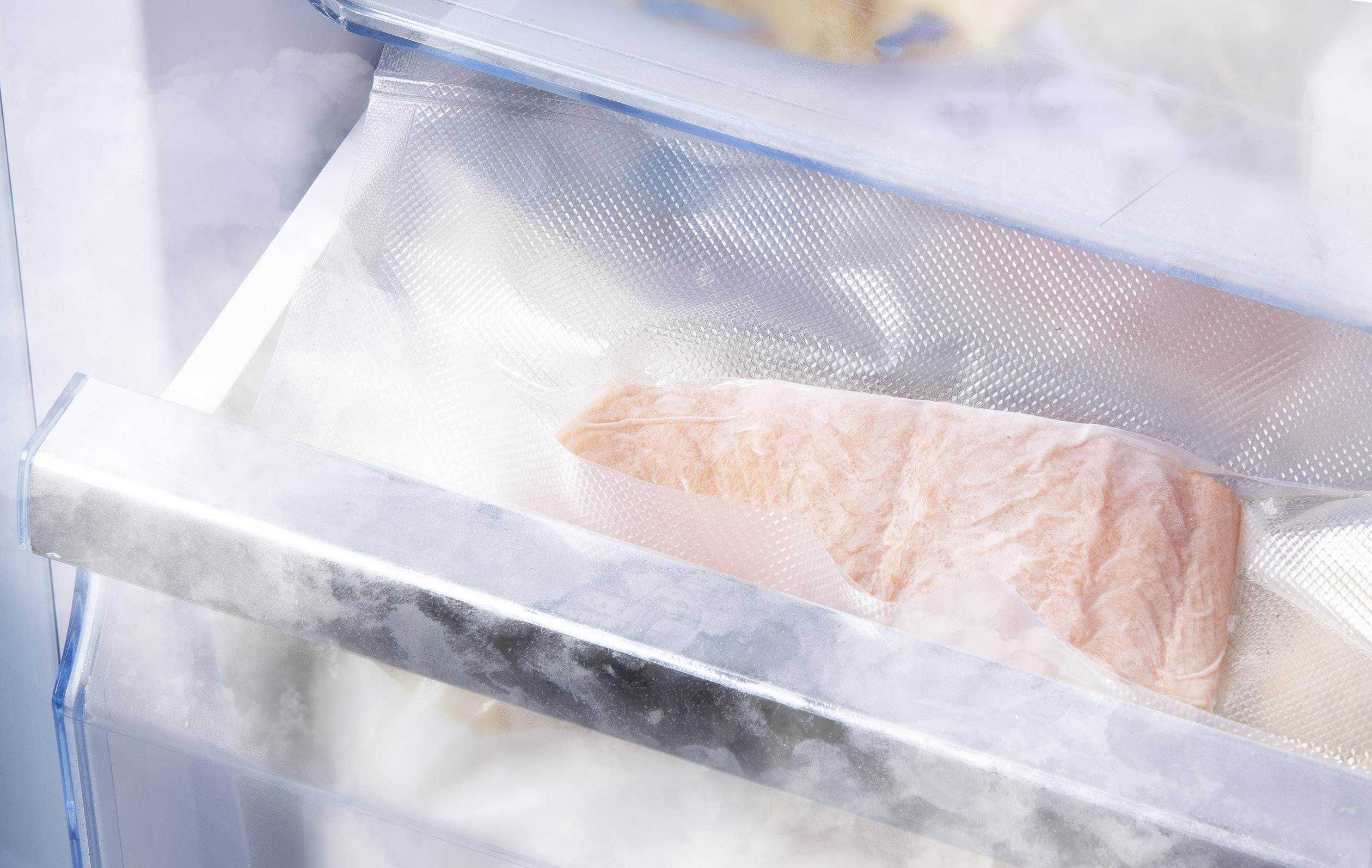 Een stuk zalm ligt vacuüm verpakt in een vriesvak. De vis is licht bedekt met rijp, wat wijst op koeling.