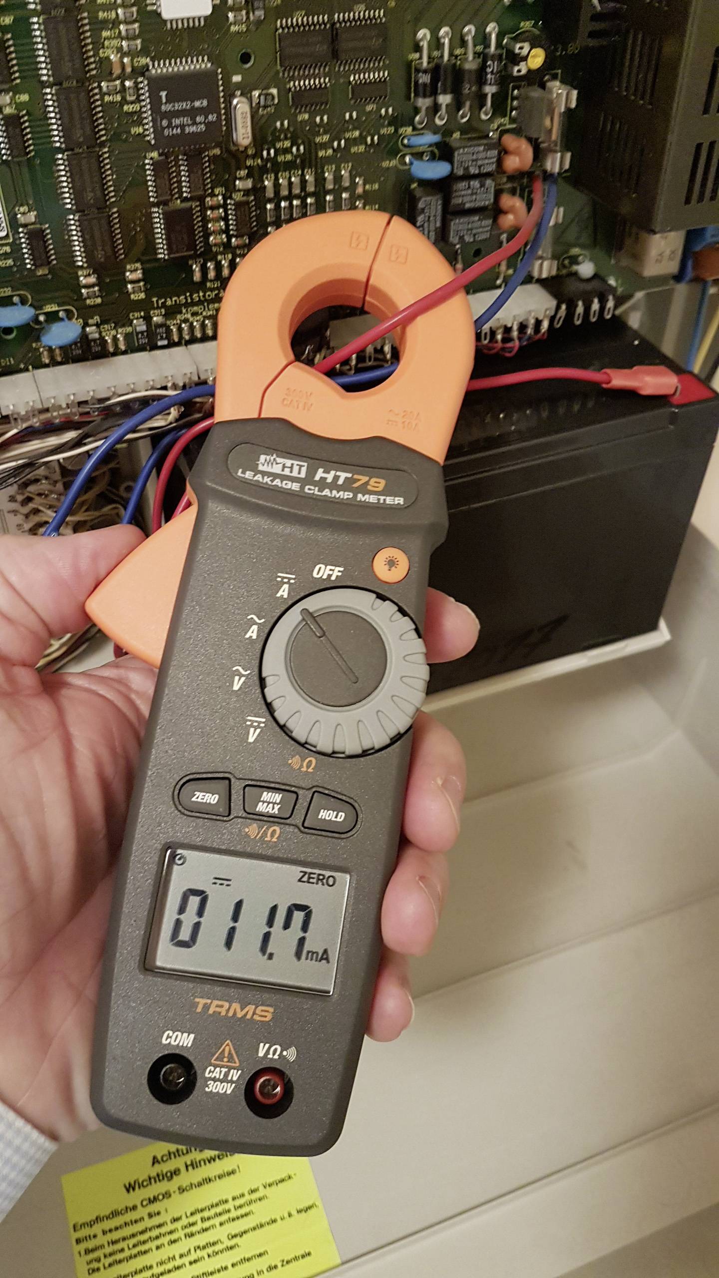 Een persoon houdt een digitale ampèretang vast die 0,117 ampère aanwijst. Op de achtergrond is een geopende elektrische schakeling zichtbaar.