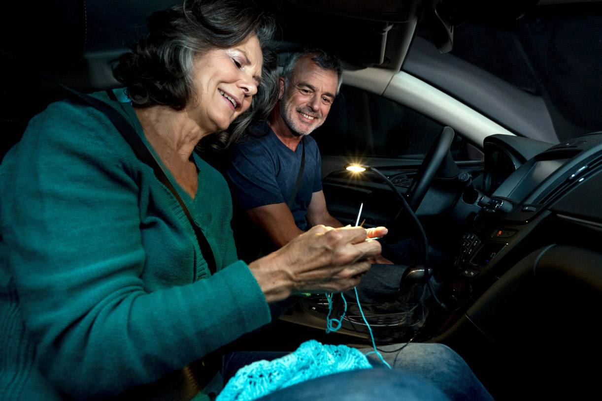 Een vrouw breit in een auto, terwijl een man haar glimlachend gadeslaat. Het is donker buiten, maar een klein lampje verlicht het interieur.