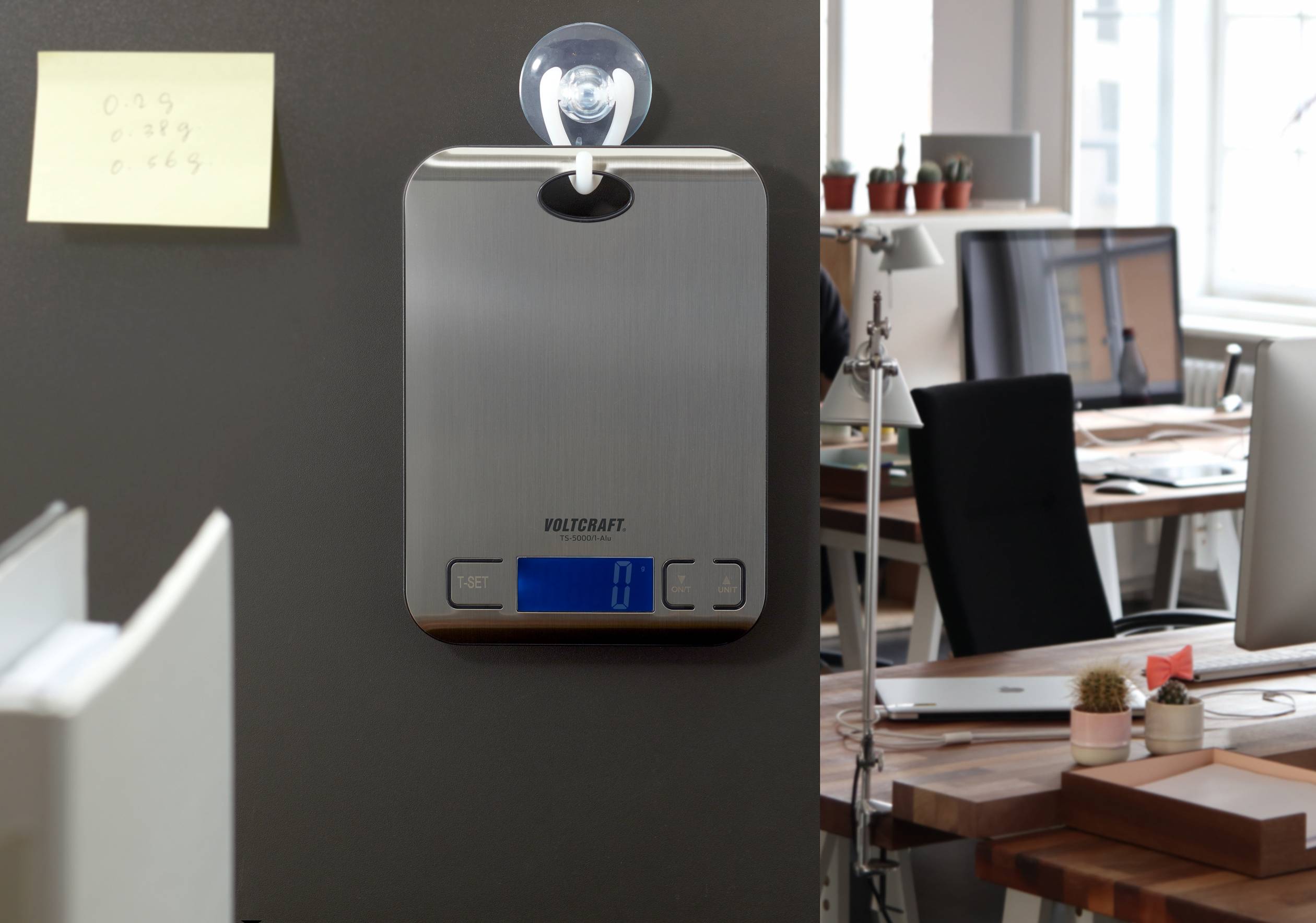 Een digitale keukenweegschaal hangt met een zuignap aan een muur in een kantoor met moderne bureaus en computers.