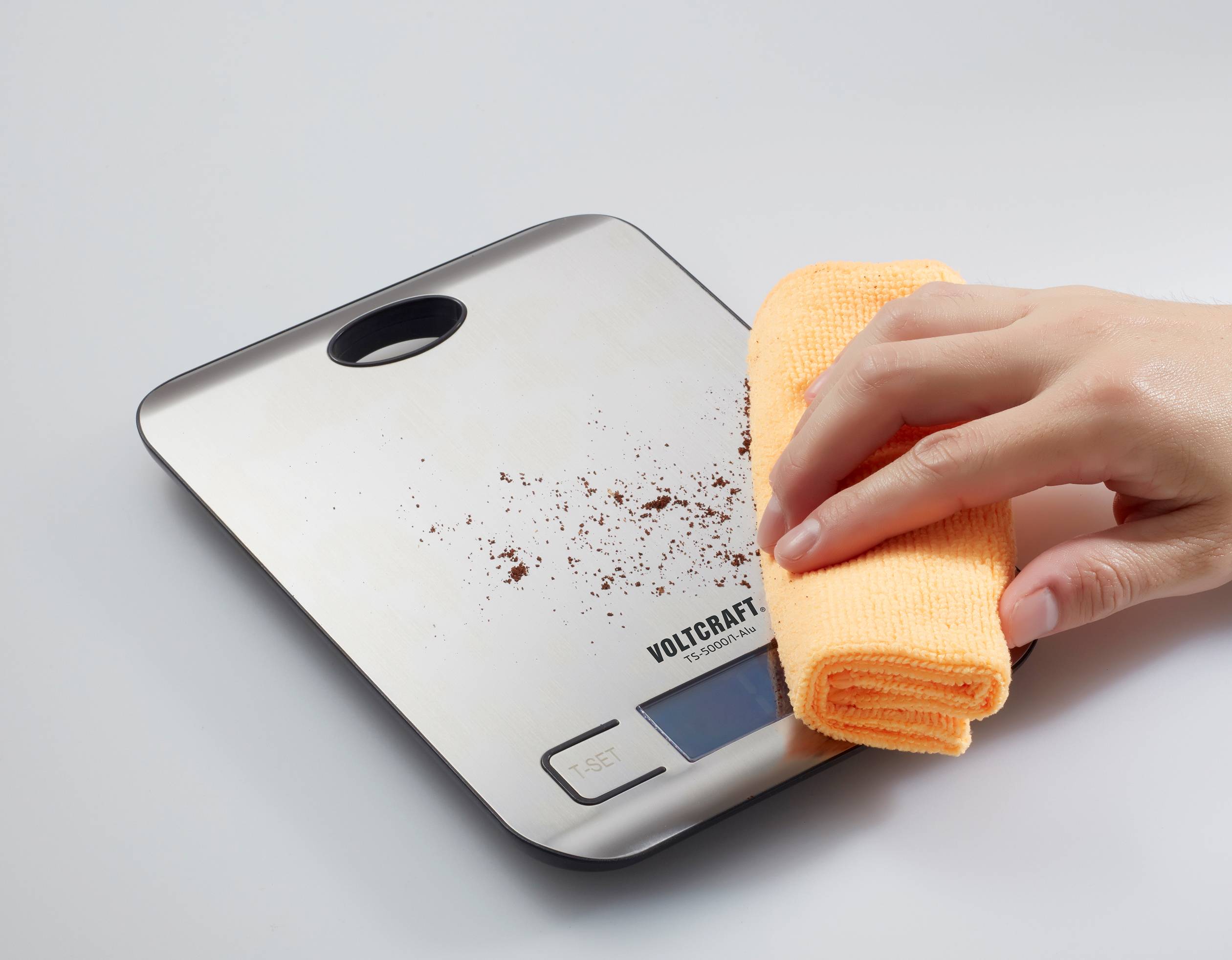 'Hand veegt koffiemeel van digitale keukenweegschaal met gele doek.'