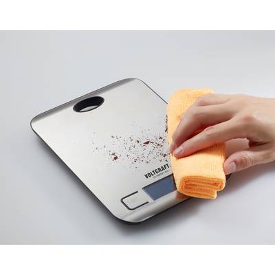 'Hand veegt koffiemeel van digitale keukenweegschaal met gele doek.'