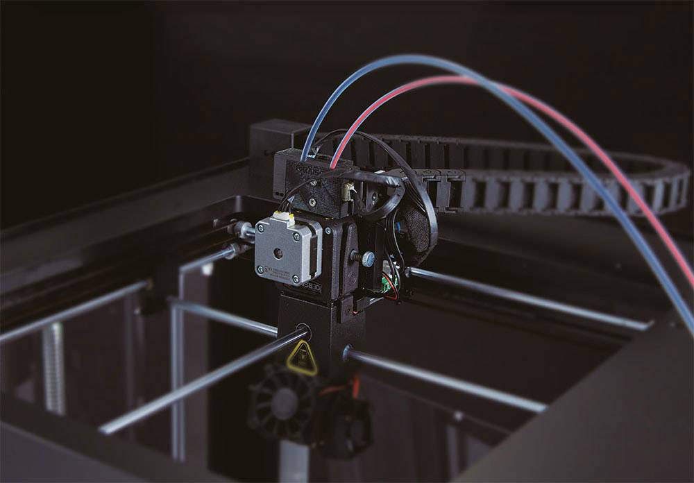 Een 3D-printer in werking drukt een object. Verschillende kabels zijn aangesloten, en de printkop beweegt zich langs de rail.