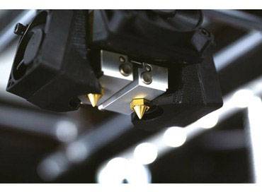 Een 3D-printer aan het werk, toont twee printkoppen met gloeiende spuitopeningen tegen een onscherpe achtergrond.