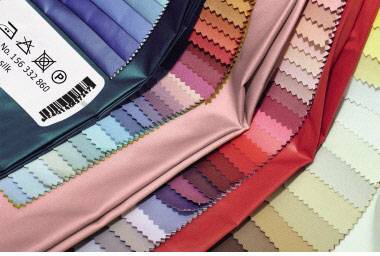 Textielstalen in verschillende kleurschakeringen van blauw, roze, rood tot beige, met verzorgingsinstructies op een etiket aan de linkerkant.