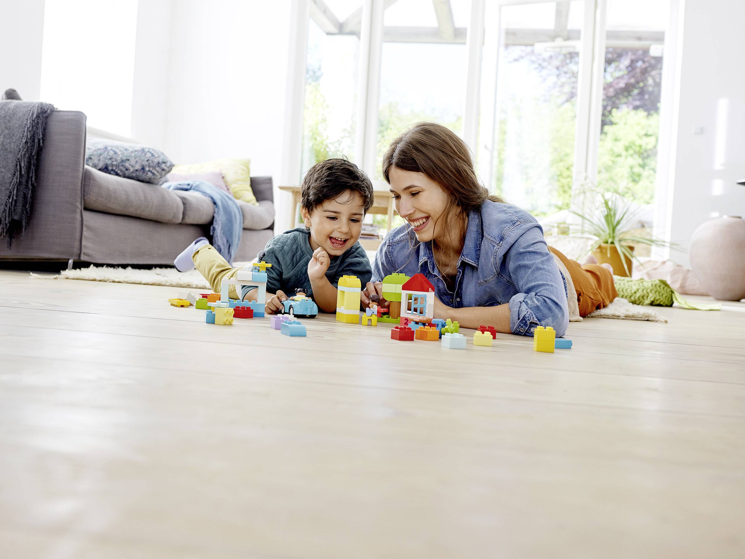 Een vrouw en een kind spelen lachend met blokken op de vloer van een lichte woonkamer.