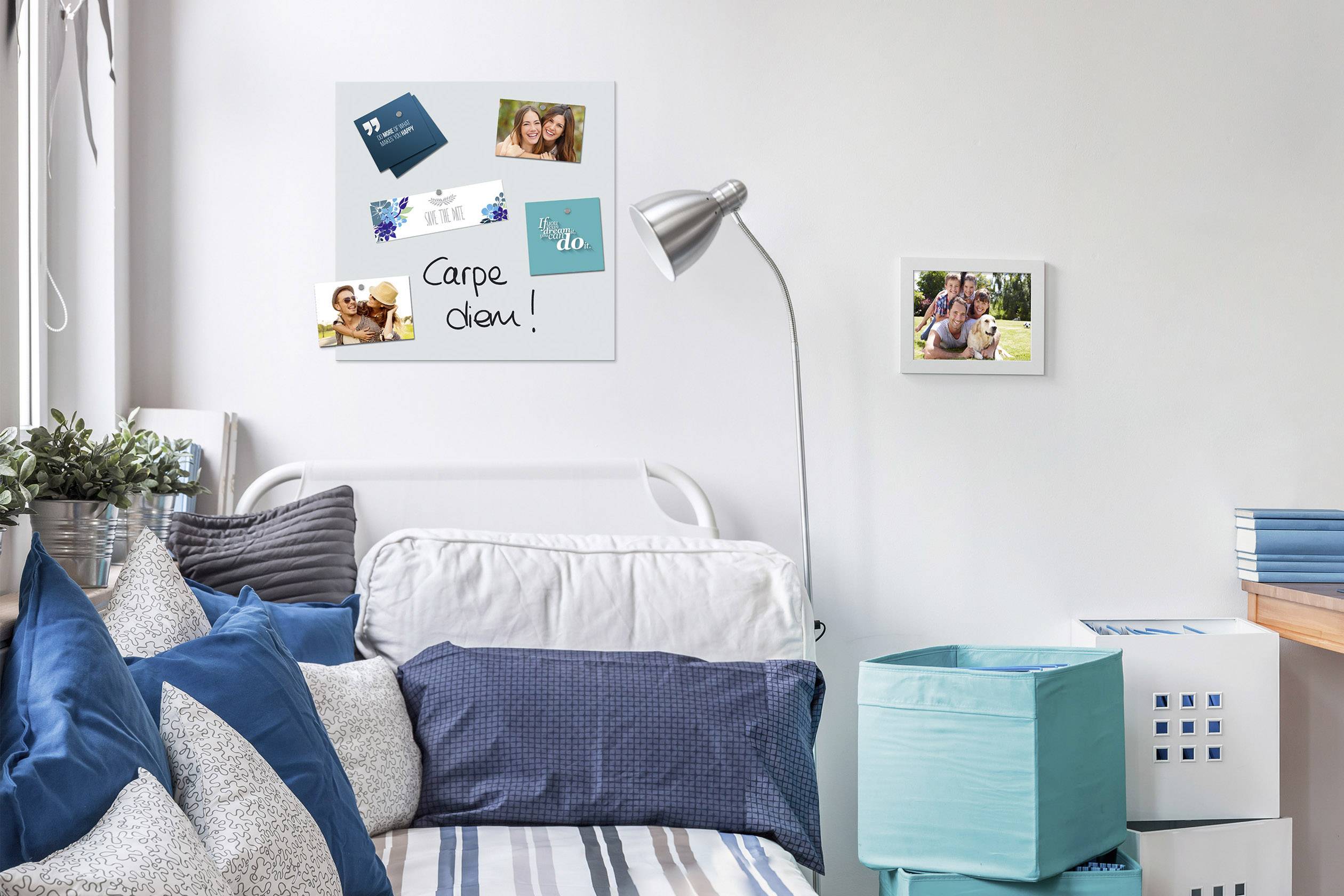 Een nette, moderne woonkamer met een bed vol blauwe kussens, een lampvoet en aan de muur een memobord met 'Carpe diem!'.