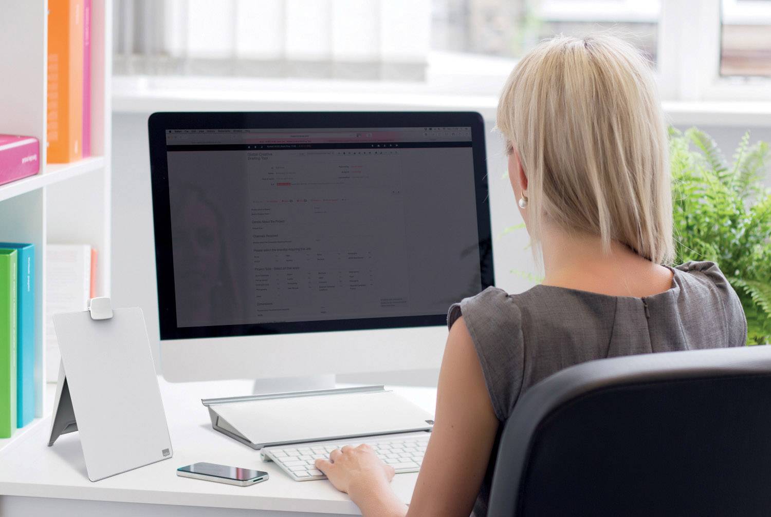 Een vrouw zit aan een bureau en werkt op een computer. Naast haar ligt een smartphone. Op de achtergrond zijn planten te zien.