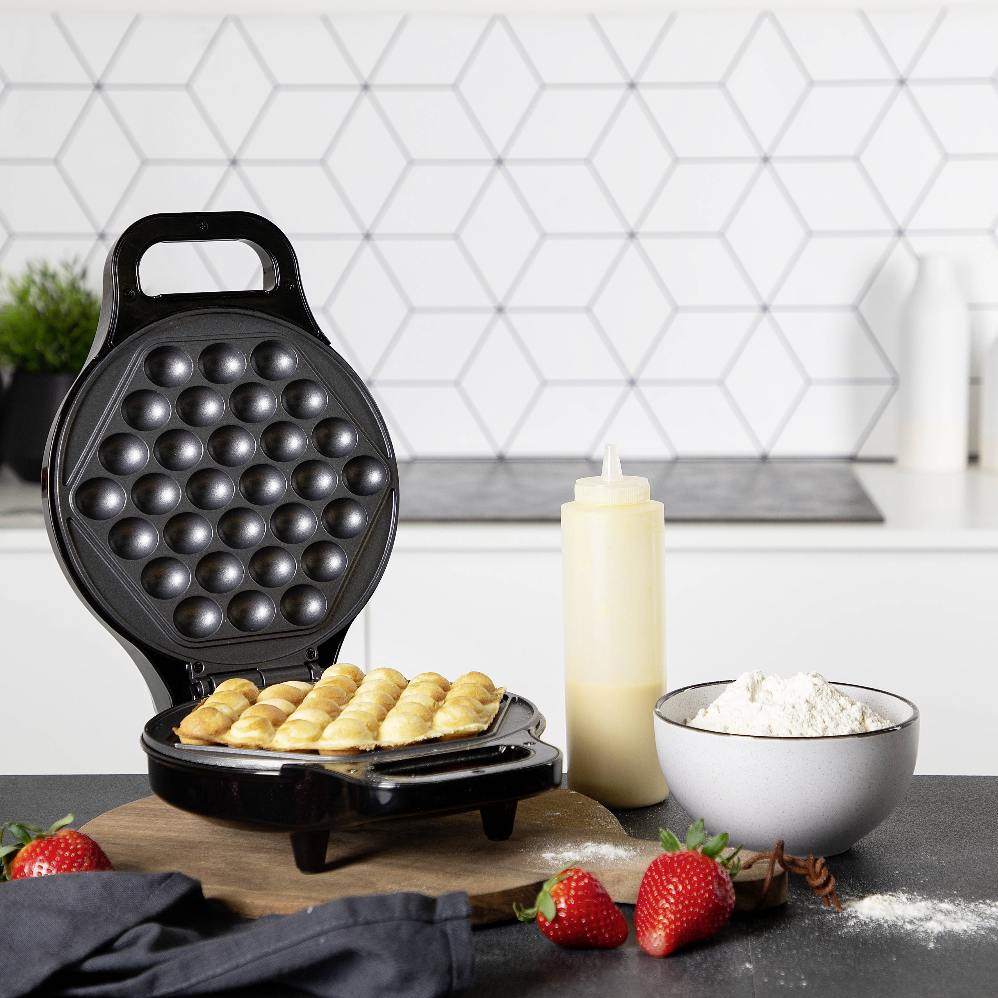 Een wafelijzer bakt eierwafels, omgeven door aardbeien, een kom meel en een knijpfles met beslag in een moderne keuken.
