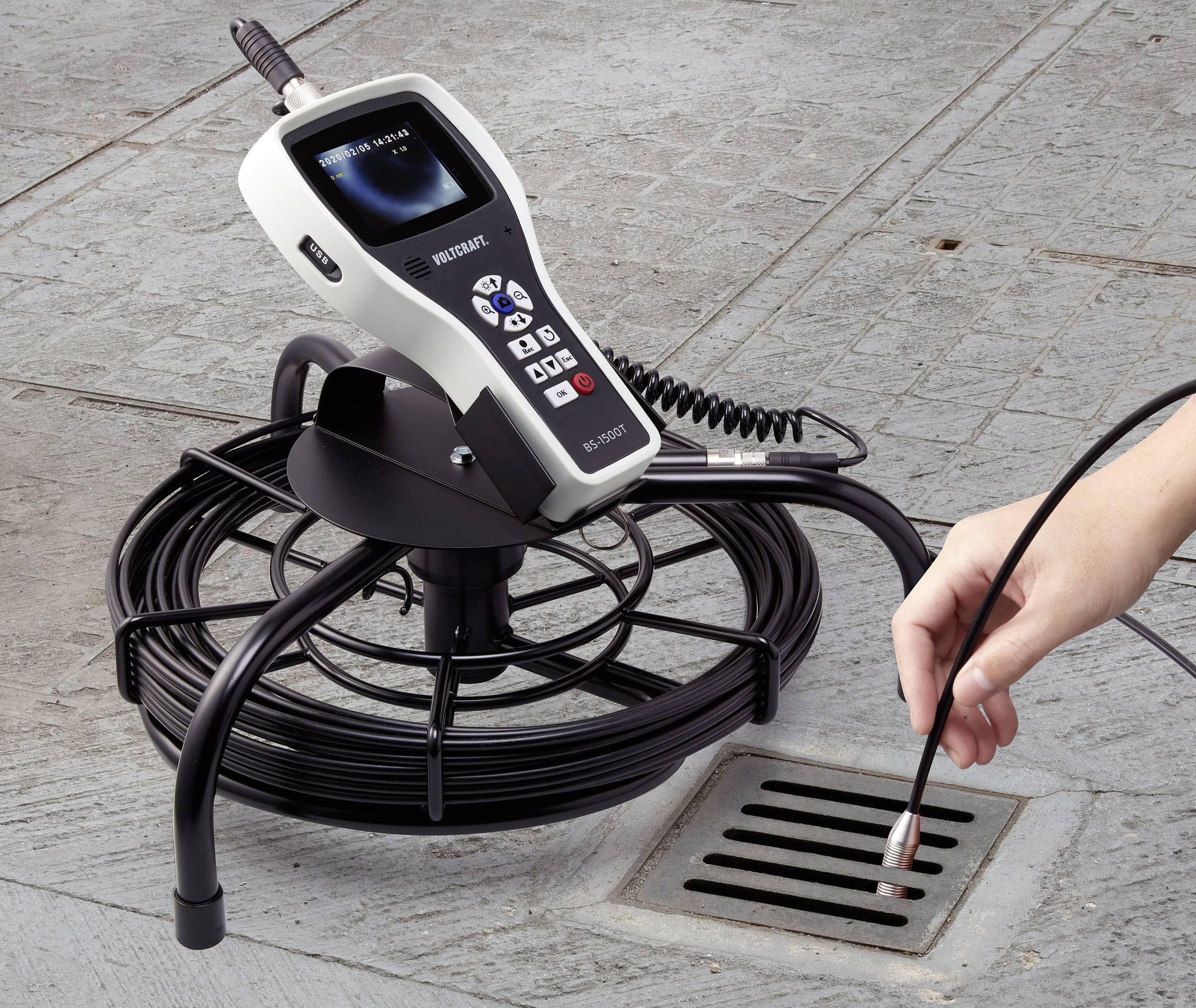 Een inspectieapparaat voor het controleren van afvoeren staat op de grond afgebeeld. Een hand brengt een camerasonde in een afvoer aan.