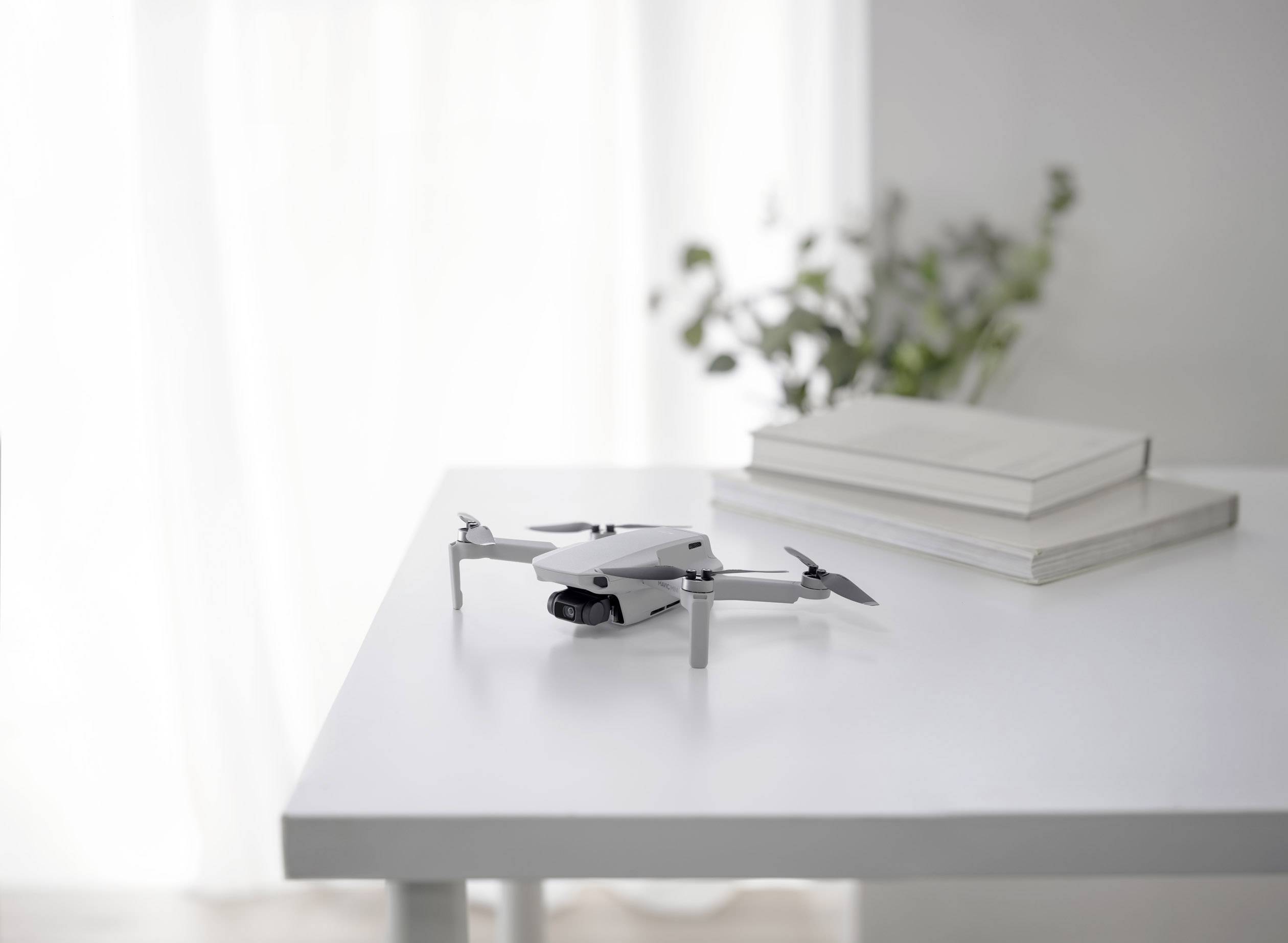 Een kleine drone ligt op een witte tafel naast twee gesloten boeken in een lichte ruimte met planten op de achtergrond.