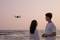Een man en een vrouw aan het strand bekijken bij zonsondergang een drone die boven de horizon zweeft.
