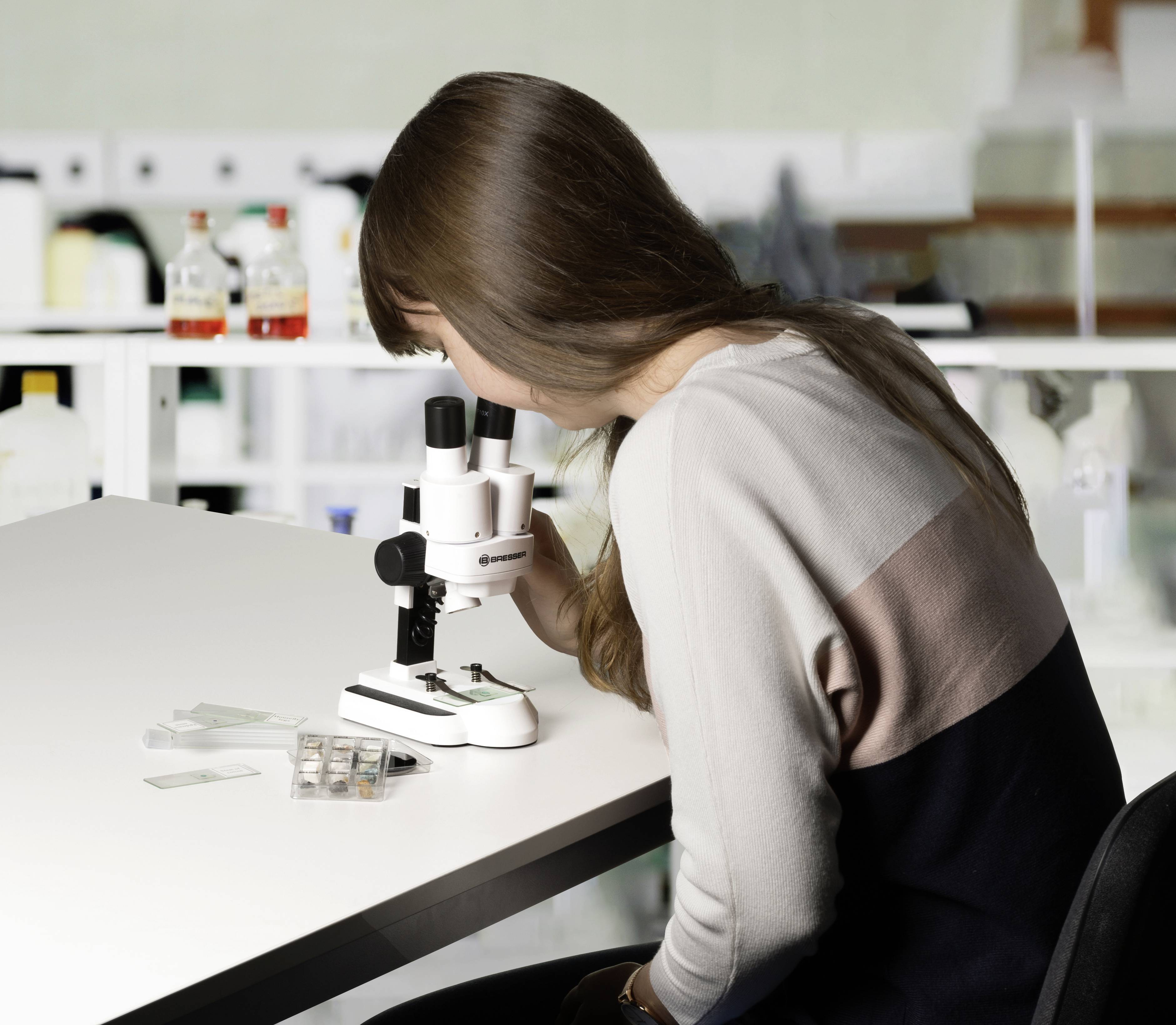 Een vrouw onderzoekt monsters in het laboratorium met een microscoop. Op de achtergrond staan chemische flessen op een rek.