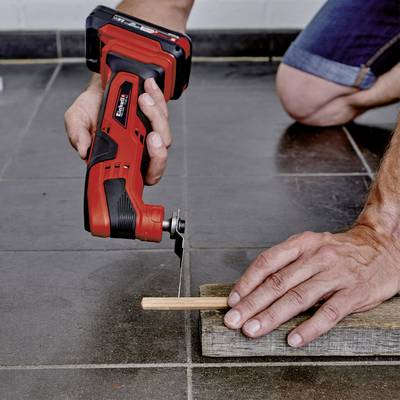 Een persoon gebruikt een rood elektrisch gereedschap om een dun stuk hout door te zagen. Het hout ligt op een grijze tegelvloer.