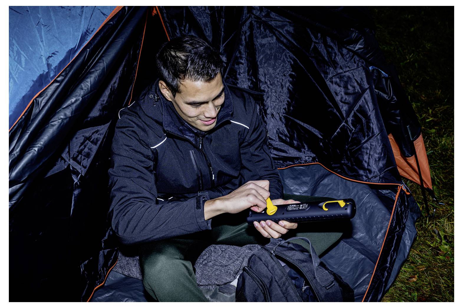 Een man in een tent gebruikt een handheld apparaat, waarschijnlijk om campingapparatuur 's nachts aan te passen of op te pompen.