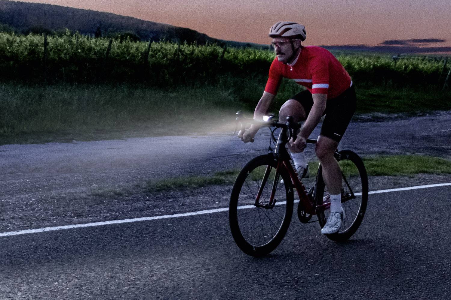 Een wielrenner met een rood shirt en helm rijdt bij schemering op een weg. Zijn fietslicht verlicht het pad voor hem. Op de achtergrond is een rij bomen zichtbaar.