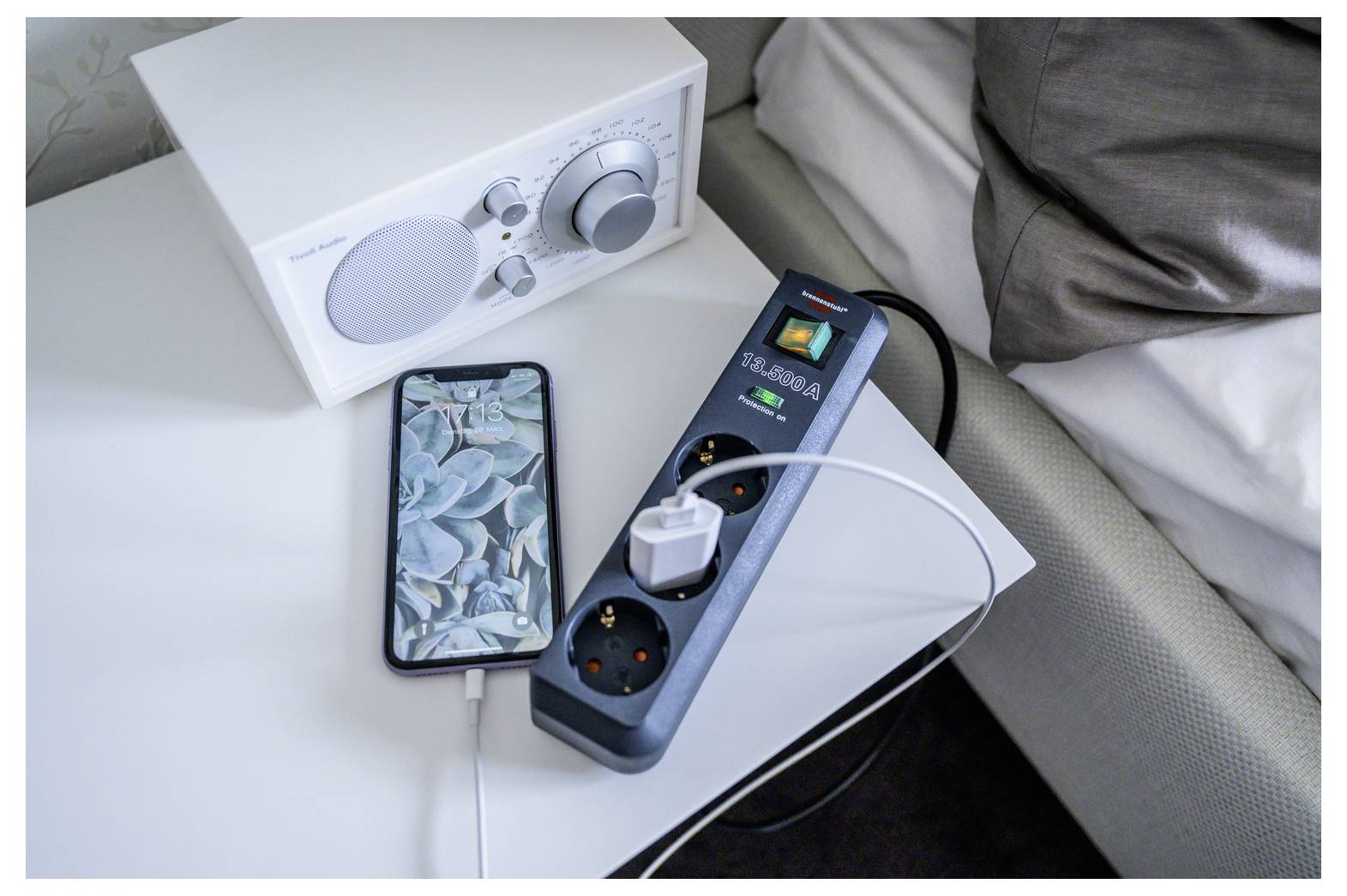 Smartphone die naast een witte speaker en een overspanningsbeveiliging met meerdere aangesloten apparaten op een nachtkastje wordt opgeladen.