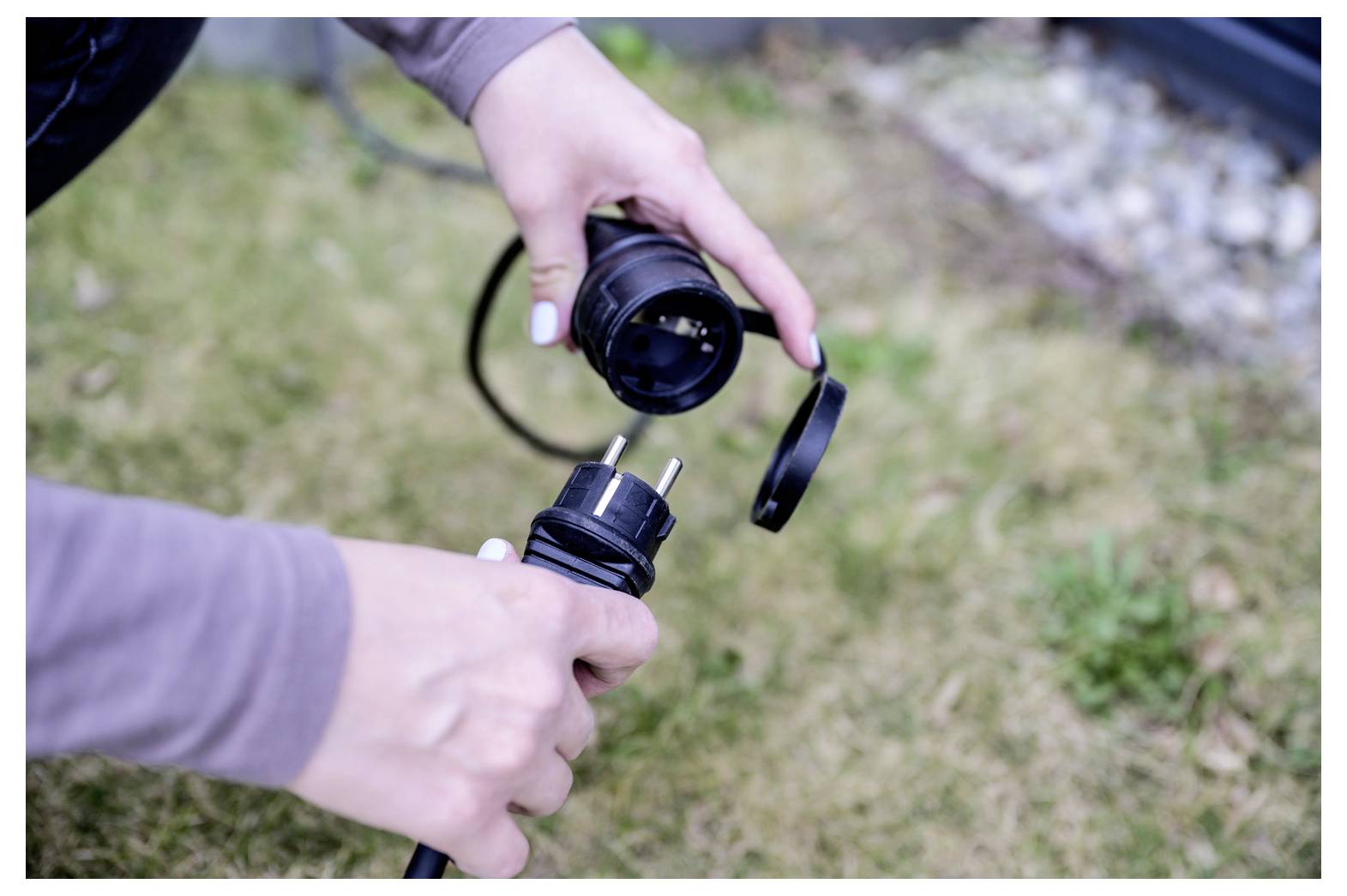 Handen die een zwarte stroomkabel met twee-polige stekker en contactdoos buiten op gras uitstekken.
