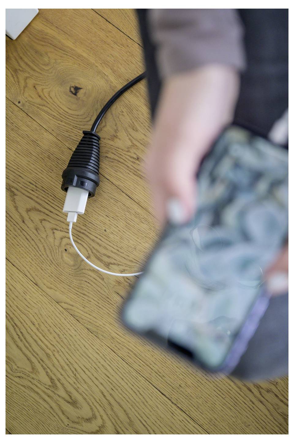 Close-up van een USB-oplaadkabel op een houten vloer terwijl een persoon een onscherp smartphone in de voorgrond vasthoudt.