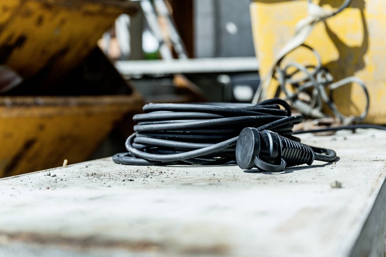 Een zwart verlengsnoer ligt op een stoffige houten tafel in een werkplaatsomgeving, met verschillende apparaten op de achtergrond.