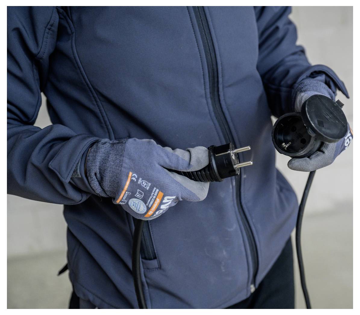 Persoon met handschoenen houdt een zwarte elektrische stekker en bijpassende contactdoos vast, waarbij een verlengkabelverbinding in voortgang wordt getoond.