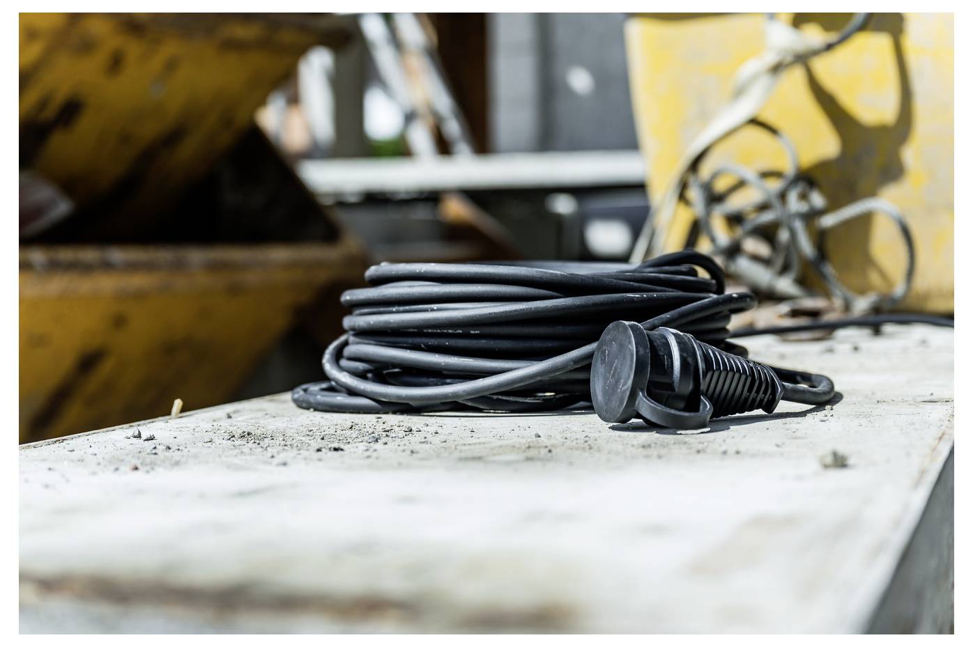 Opgerolde zwarte elektrische kabel met stekker rustend op een stoffig werkblad in een industriële werkplaats.