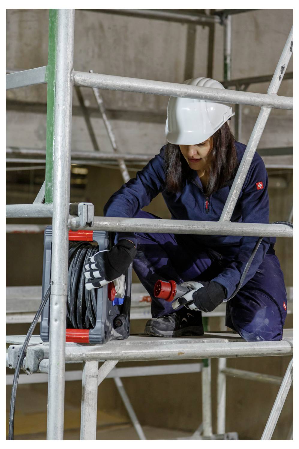 Bouwvakker in een helm en handschoenen gebruikt een elektrische boor terwijl hij gehurkt op steigers in een gebouw in aanbouw staat.
