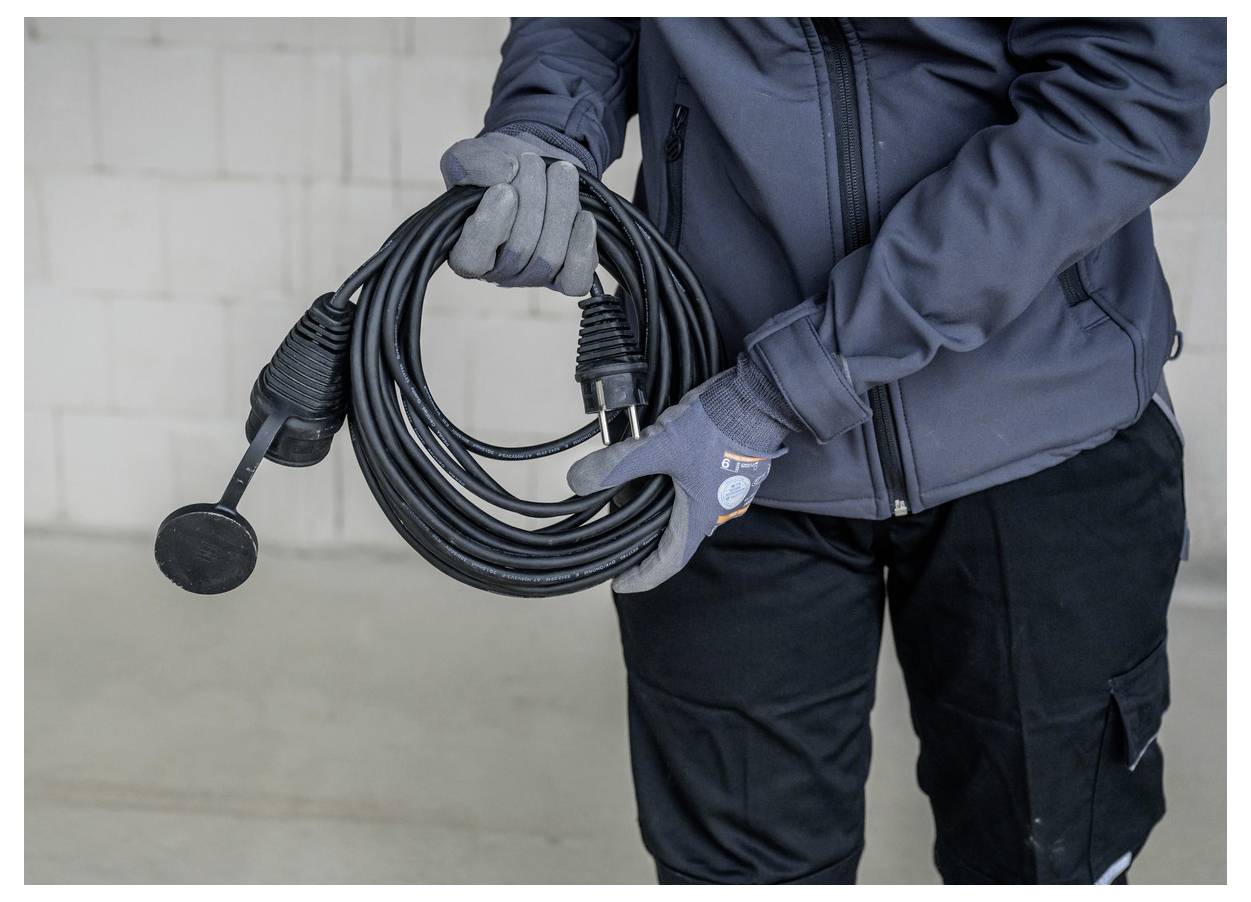 Gehandschoende werker die een opgerold zwart kabel met een stekker en microfoonachtige connector tegen een gewone binnenmuur vasthoudt.