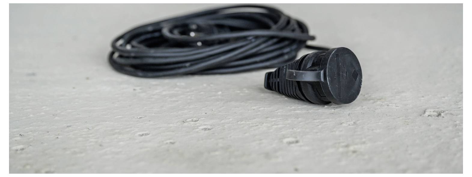 Close-up van een zwarte elektrische snoer of kabel met een stekker die opgerold op een lichte grijze betonnen vloer ligt.