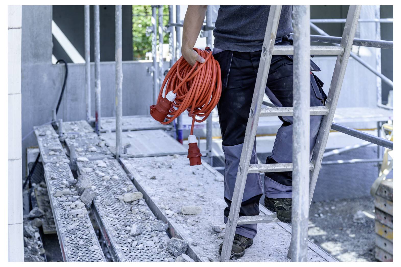 Bouwvakker die een oranje verlengsnoer vasthoudt terwijl hij op steigers staat naast een ladder op een bouwplaats.