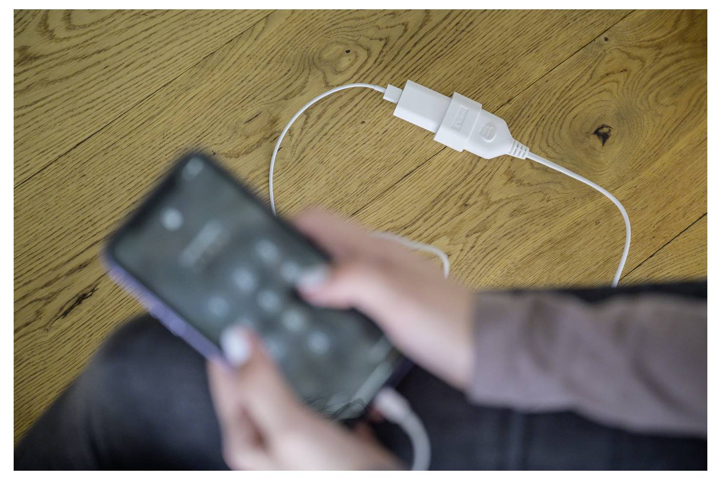 Wazig persoon die een smartphone gebruikt die via een witte USB-kabel is aangesloten op een stromaadapter op een houten vloer.
