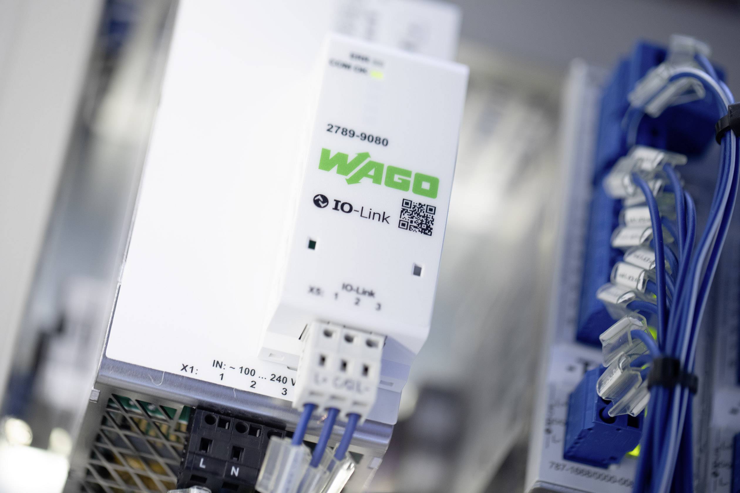 Een IO-Link-apparaat van een elektronicafabrikant staat centraal. Naast het apparaat zijn kabels en een deel van een besturingssysteem zichtbaar.
