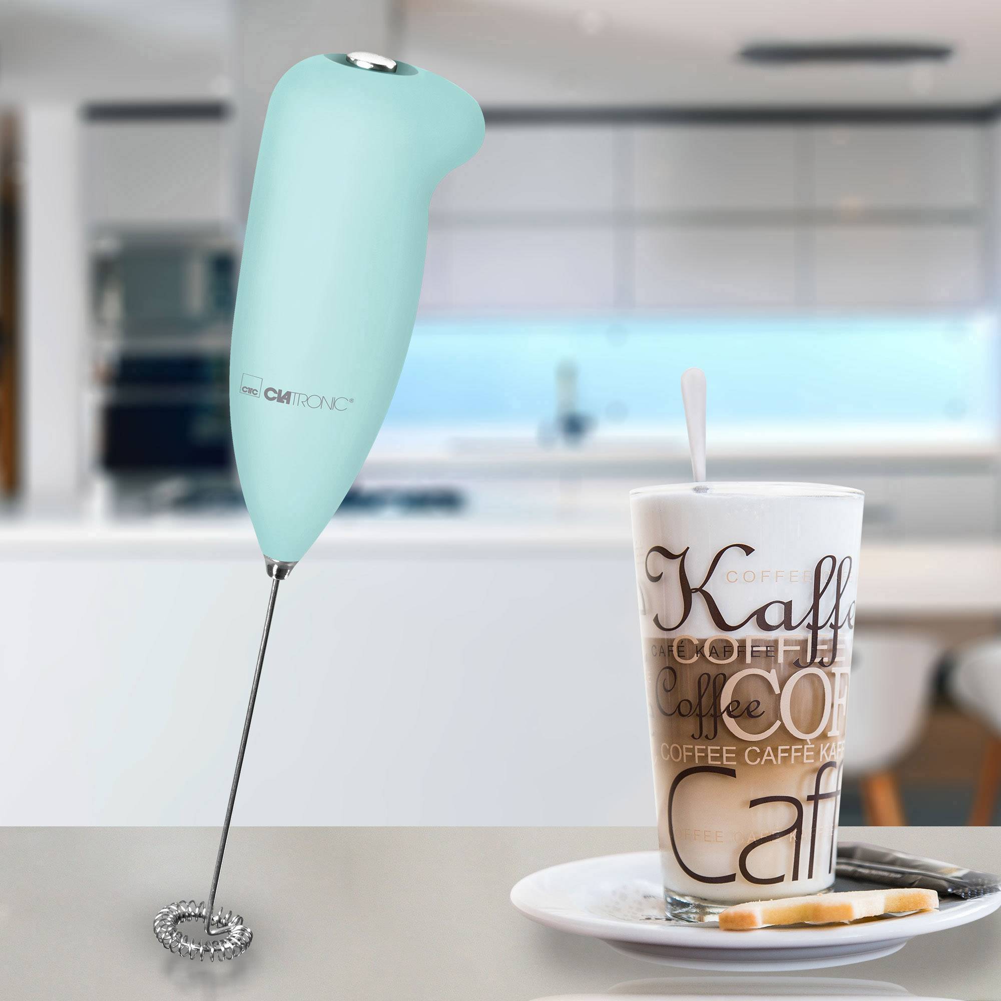 Een melkopschuimer naast een glas koffie in een moderne keuken. De opschuimer is turquoise van kleur en staat rechtop.