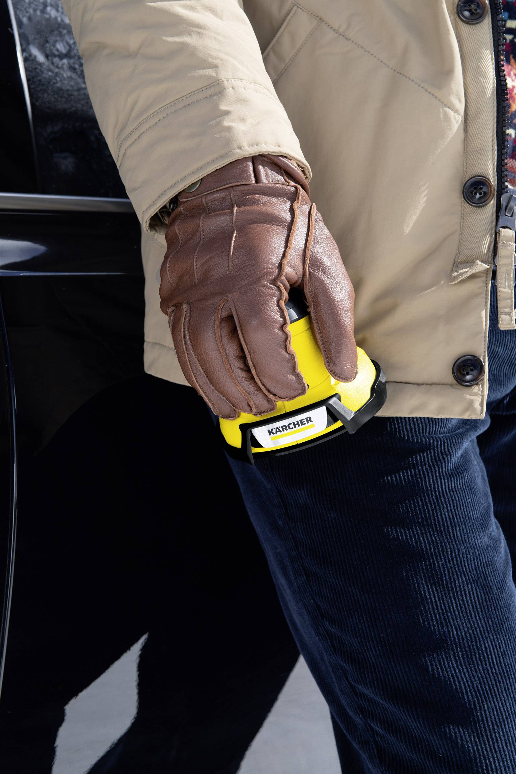 Een persoon draagt bruine handschoenen en houdt een geel apparaat met het opschrift 'Kärcher' naast een zwarte auto.