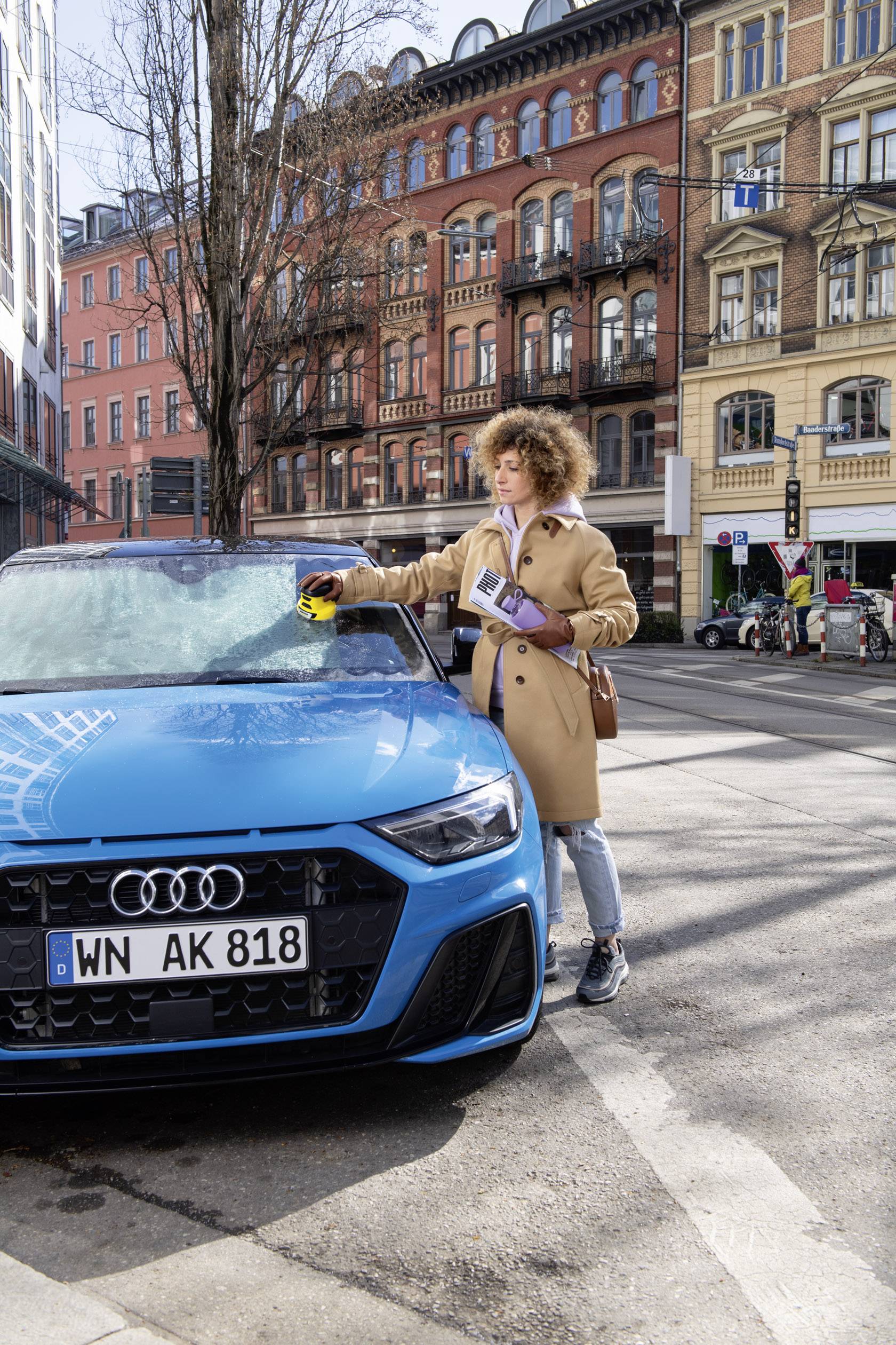 Een persoon verwijdert ijs van de voorruit van een blauwe auto met een ijskrabber. Op de achtergrond zijn gebouwen van een stad zichtbaar.