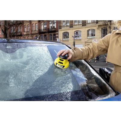 Een persoon verwijdert ijs van de voorruit van een auto met een elektrisch apparaat. Op de achtergrond zijn gebouwen zichtbaar.