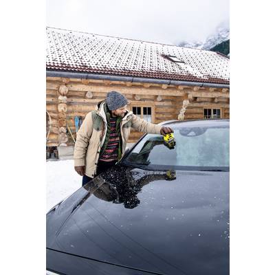 Een persoon in winterkleding schraapt ijs van de voorruit van een auto voor een houten huis in de sneeuw.