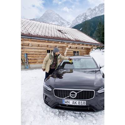 Een man in winterkleding veegt sneeuw van een zwarte auto voor een houten hut en een met sneeuw bedekt berglandschap.