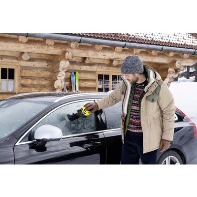 Een man krabt ijs van de autoruit in een besneeuwde omgeving voor een blokhut. Ski's staan voor de hut.